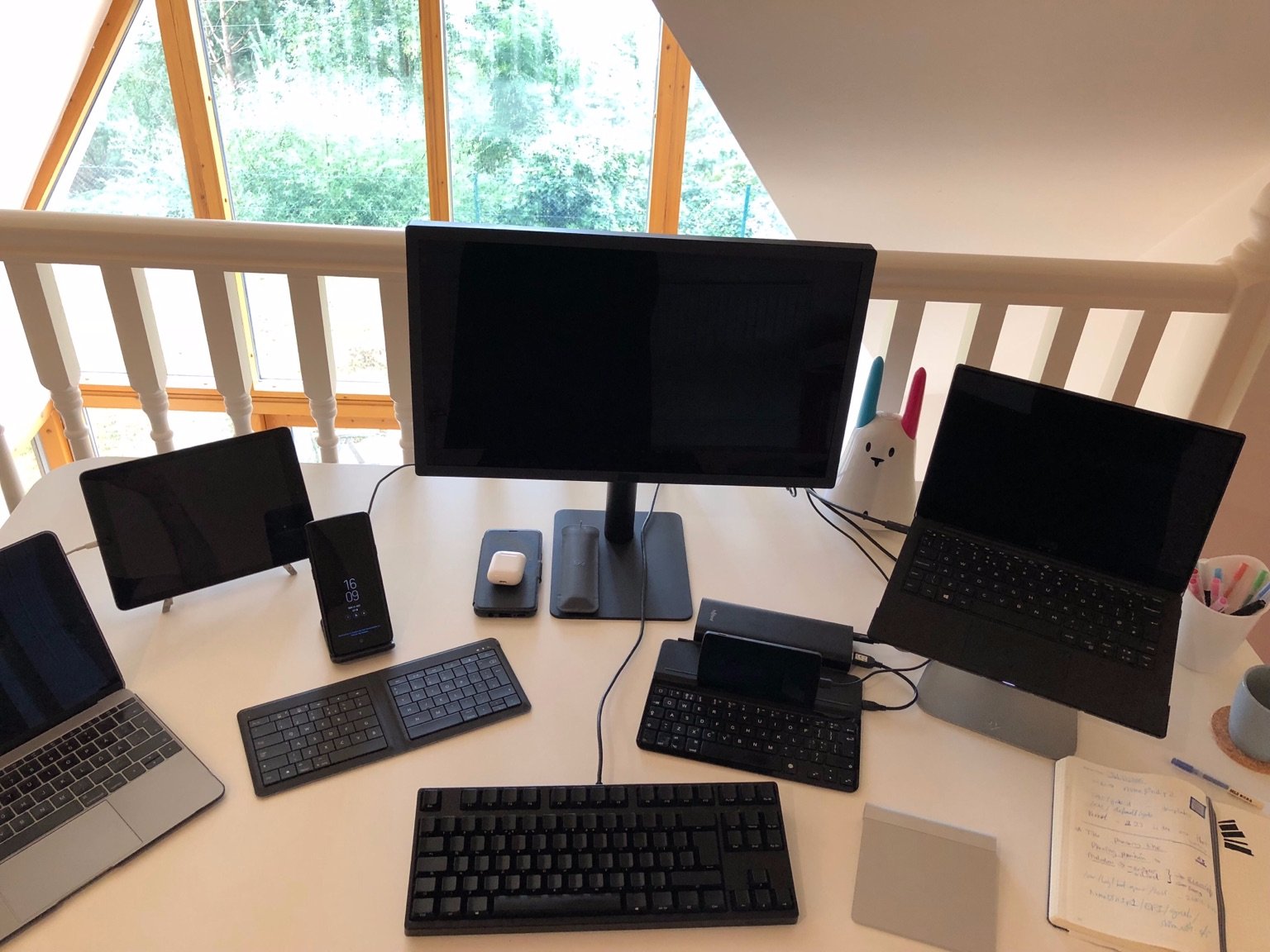 Photo of my desk showing a number of devices: from left to right, my MacBook, and iPad, a Samsung S9+ with a Microsoft foldable keyboard, a 21" LG monitor, a Filco Majestouch keyboard, a pair of AirPods in their case, a Logitech presentation remote in its gray leather sleeve, a Nexus 5 running LineageOS resting on a Logitech bluetooth keyboard originally for the iPad, a Magic Trackpad, and a Dell XPS 13 running Pop!_OS 18.04. All screens are off. Peeking from behind them is my Nabaztag bunny with one blue ear and one pink ear. To the far right, partially in the photo, is a pen holder with Muji pens, a coffee  mug, and my Leuchtturm notebook.