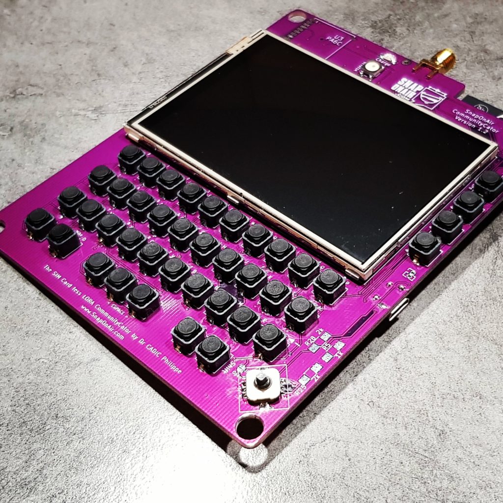 The SnapOnAir Lora communicator circuit board, purple, with buttons and a screen. Unit is off.
